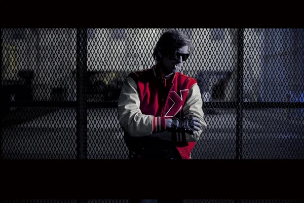 Kavinsky, le DJ français, en veste de varsity rouge et blanche et lunettes de soleil, bras croisés devant un grillage, image encadrée de barres noires évoquant un extrait de clip vidéo