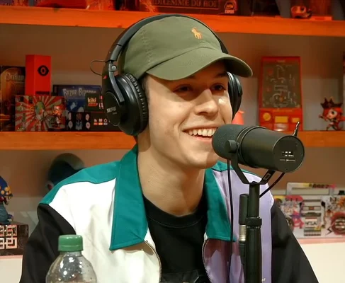Seb La Frite, casquette verte, casque audio, veste colorée, souriant et parlant dans un micro, entouré de jeux de société en studio.