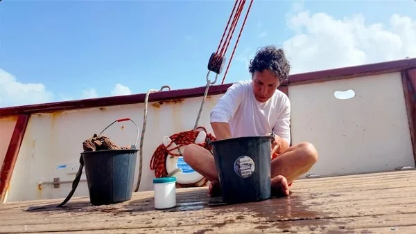Sélim Ben Hassen, cheveux bouclés et t-shirt blanc, assis en tailleur sur le pont en bois d'un bateau, lavant des objets dans un seau noir sous un ciel bleu éclatant lors de sa traversée de l'Atlantique.