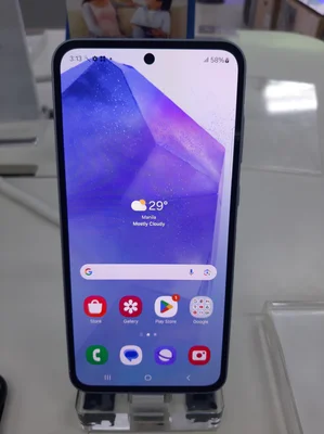 Photographie en gros plan d'un smartphone Samsung Galaxy A55 5G posé sur un présentoir en acrylique transparent, affichant l'écran d'accueil avec des icônes d'applications et un widget météo.