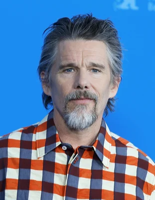 Portrait d'Ethan Hawke en chemise à carreaux orange, bleue et blanche, photographié à la Berlinale 2025, arborant cheveux grisonnants et bouc.