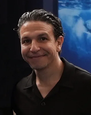 Portrait de Sélim Ben Hassen, les cheveux bruns gominés en arrière, vêtu d'une chemise noire à col, souriant légèrement devant un fond partagé entre une zone sombre et un écran bleu.