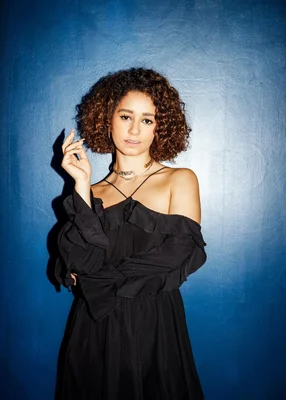 Léna Situations, cheveux bruns bouclés, robe noire décolletée et colliers superposés, posant devant un mur bleu texturé.