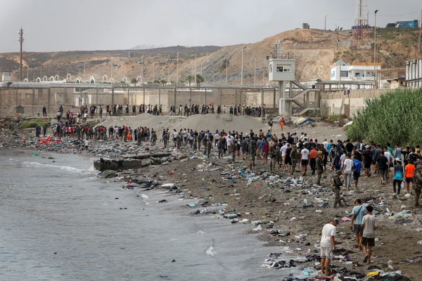 Foule de migrants et de forces de l'ordre sur une plage jonchée de débris, devant la clôture frontalière fortifiée de Ceuta avec ses miradors, lors d'une tentative de passage massif