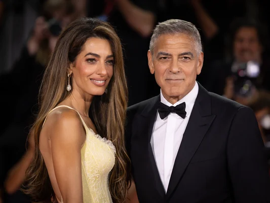 George Clooney en smoking noir et Amal Clooney en robe de dentelle jaune posant ensemble au Festival de Venise 2024, souriant à l'objectif.