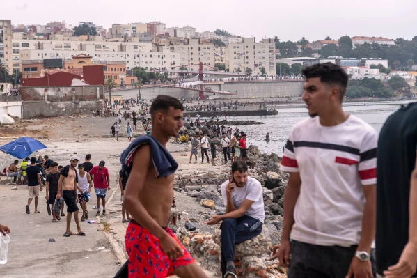 Plage bondée à Ceuta avec des migrants, dont un jeune homme torse nu en short rouge, marchant près du rivage, bâtiments côtiers et jetée en pierre en arrière-plan