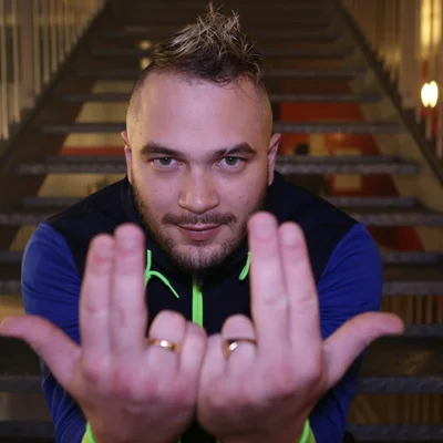 Jul, le rappeur marseillais, posant sur un escalier avec une coupe mulet et une barbe, arborant des bagues en or et une veste de survêtement bleu et noir à fermeture éclair néon verte, dans le cadre de son portrait pour ses 10 ans de carrière.
