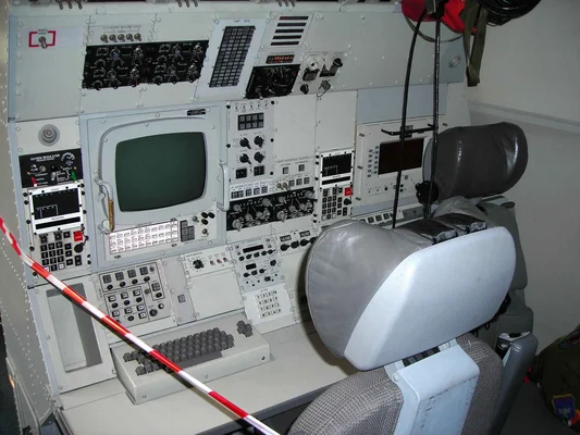 Intérieur d'un avion E-3 Sentry AWACS montrant un poste de pilotage avec siège, commutateurs analogiques, écrans numériques et panneaux de contrôle.