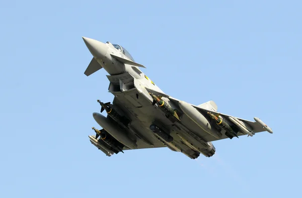 Un Eurofighter Typhoon de la Royal Air Force en vol, incliné, montrant ses missiles et réservoirs de carburant externes, sous un ciel bleu clair.