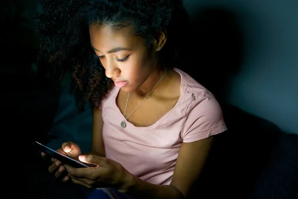 Une jeune femme aux cheveux bouclés bruns, vêtue d'un t-shirt rose, regarde son smartphone dans une pièce faiblement éclairée, le visage illuminé par l'écran.