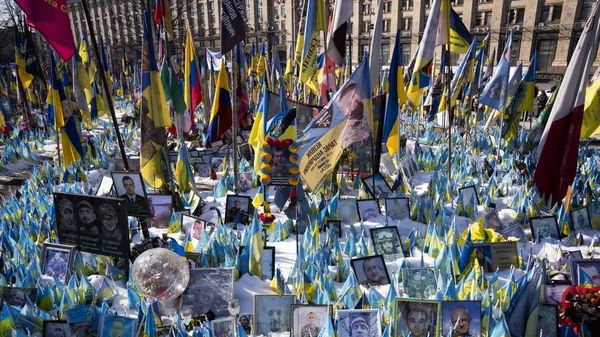 Place enneigée à Kiev avec des centaines de drapeaux ukrainiens et des portraits encadrés de soldats tombés ou de victimes, disposés au sol lors d'une journée de commémoration.