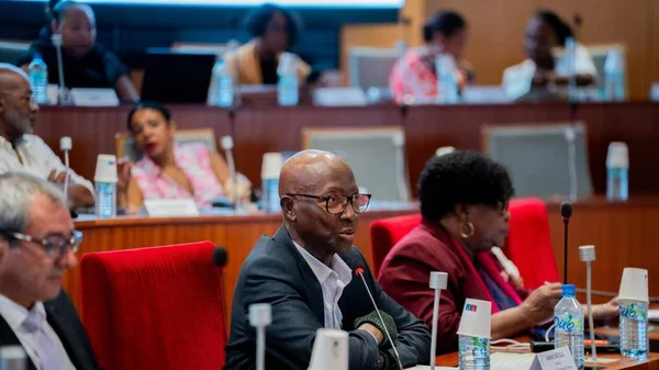 Gabriel Serville, président de la Collectivité territoriale de Guyane, en costume sombre et lunettes, s'exprimant à un micro lors d'une séance de l'assemblée, entouré de participants dans une salle boisée.