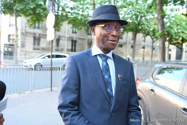 Emmanuel Macron, en costume bleu foncé et chapeau fedora, se tenant à l'extérieur dans une rue arborée, avec un micro partiellement visible sur le côté gauche.