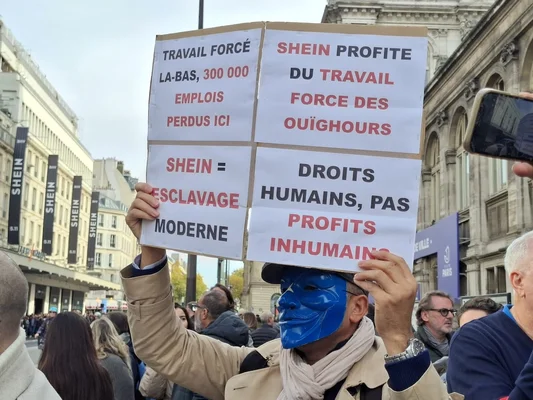 Un manifestant portant un masque bleu tient une pancarte en français critiquant Shein pour le travail forcé et les violations des droits humains, devant un bâtiment affichant des bannières Shein.