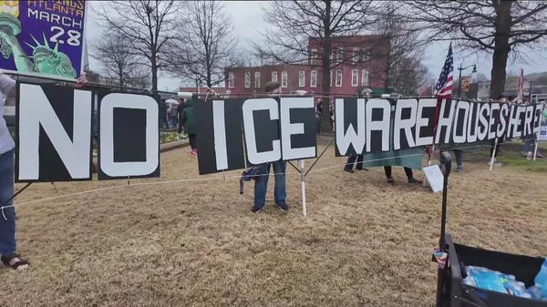 Manifestation à Social Circle, en Géorgie, où des habitants brandissent des pancartes blanches sur lesquelles on peut lire « NO ICE WAREHOUSE HERE » devant un bâtiment en briques rouges, pour protester contre le futur centre de détention d'ICE.