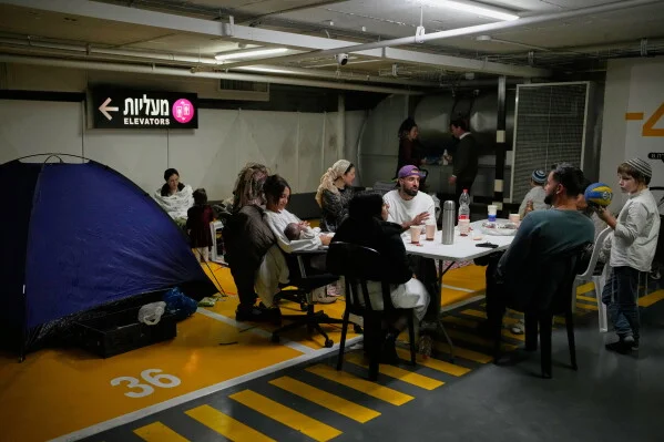 Des familles israéliennes, dont des enfants et un bébé, se réfugient dans un parking souterrain transformé en abri lors d'une attaque de missiles iraniens, avec une tente bleue et des panneaux en hébreu visibles sur le mur.