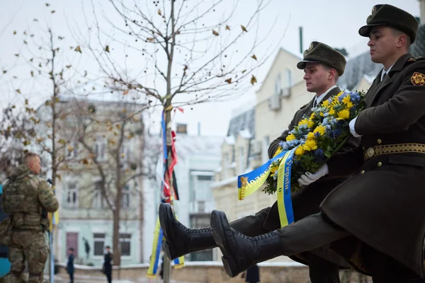 Deux soldats en uniforme sombre et calot portant une couronne de fleurs aux rubans bleu et jaune lors d'une cérémonie commémorative, avec un soldat en camouflage en arrière-plan près du Mur du Souvenir à Kyiv.