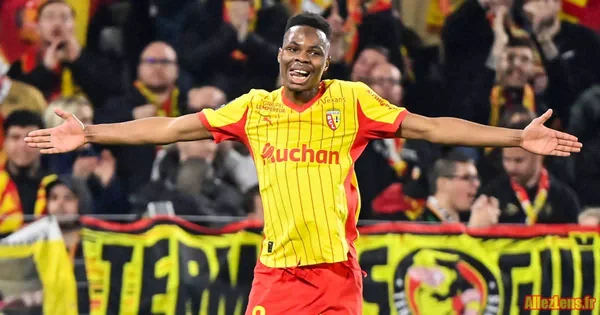 Mamadou Sangaré, joueur de football en maillot rayé jaune et rouge floqué Auchan, célébrant sur la pelouse, les bras écartés, devant des supporters flous aux couleurs du RC Lens.