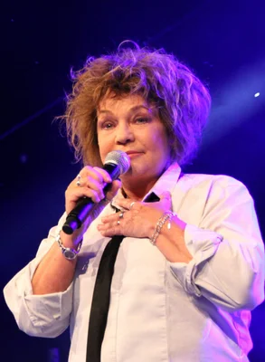 Sabine Paturel sur scène, cheveux courts et bouclés, chemise blanche et cravate noire, chantant dans un microphone sous un éclairage violet.