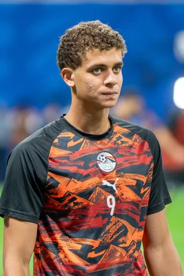 Hamza Abdelkarim, jeune footballeur égyptien aux cheveux bouclés, en maillot d'entraînement orange et noir portant le numéro 9, dans un stade flou en arrière-plan.