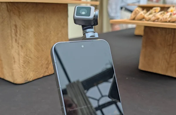 Gros plan sur un smartphone Honor avec une caméra d'action carrée fixée sur sa tranche supérieure via un adaptateur, l'écran sombre reflétant l'environnement intérieur avec une fenêtre et une structure de plafond
