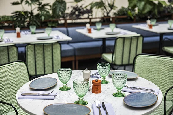 Table ronde en marbre du restaurant Montecito au Kimpton Saint-Honoré à Paris, dressée avec des verres verts texturés et des assiettes bleues, avec en arrière-plan des banquettes à motifs et un mur de plantes d'intérieur