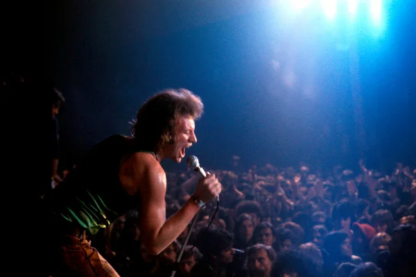 Johnny Hallyday, cheveux bruns ébouriffés et débardeur noir, chantant intensément dans un micro sous des projecteurs bleus lors d'un concert, avec une foule nombreuse en arrière-plan.