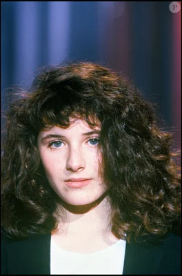 Elsa Lunghini en 1986, jeune chanteuse aux cheveux bruns bouclés et aux yeux bleus, veste noire sur haut blanc, fond dégradé bleu et violet.