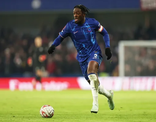 Axel Disasi en maillot bleu de Chelsea dribblant le ballon sur la pelouse lors d'un match, avec les lumières du stade et des spectateurs flous en arrière-plan.