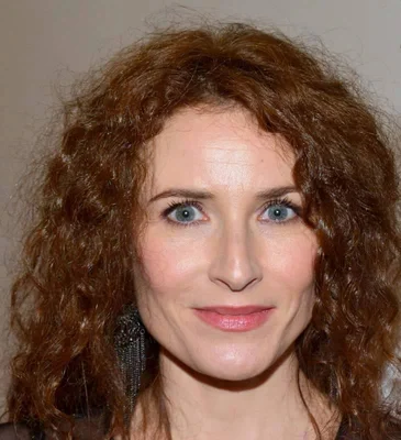 Portrait récent d'Elsa Lunghini, cheveux bouclés auburn et yeux bleus, souriant légèrement face à l'objectif.