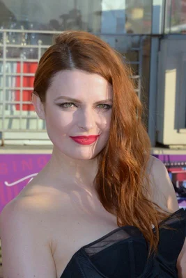 Élodie Frégé, gagnante de la Star Academy, souriant avec ses cheveux roux ondulés et un rouge à lèvres vif, vêtue d'une robe noire sans bretelles devant un fond violet flou.