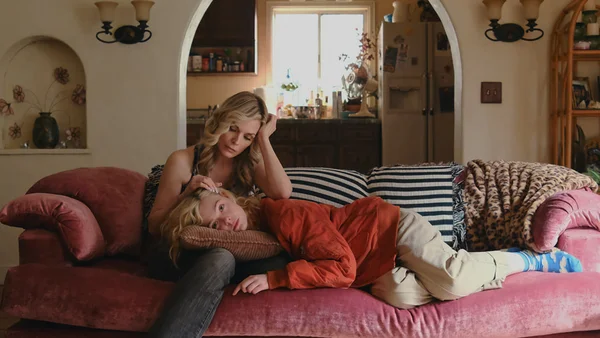 Michelle Pfeiffer assise sur un canapé en velours rose, touchant le front d'une jeune fille en veste orange, dans un salon aux arches donnant sur une cuisine