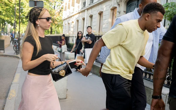 Kylian Mbappé et Ester Expósito lors d'une virée shopping à Paris, photographiés dans la rue, elle en haut noir et jupe rose tenant son téléphone, lui en t-shirt jaune, avec des passants en arrière-plan.