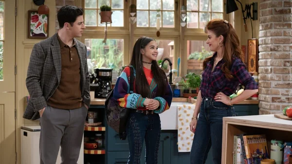 David Henrie et Janice LeAnn Brown dans une scène de la saison 2 de Wizards Beyond Waverly Place, debout dans une cuisine avec une autre actrice, portant des chemises à carreaux et un pull coloré avec sac à dos.