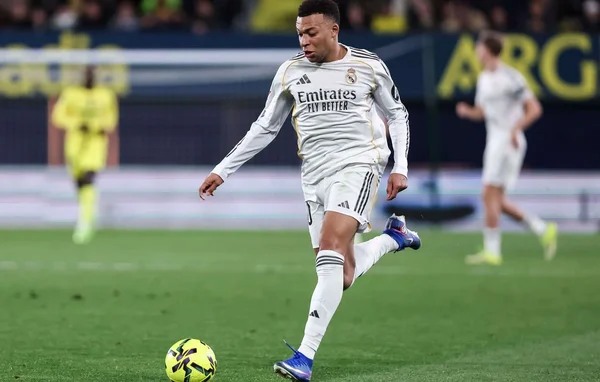 Kylian Mbappé en maillot blanc du Real Madrid dribblant un ballon jaune sur la pelouse verte lors d'un match de Liga, avec un arrière-plan flou montrant le stade et d'autres joueurs