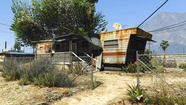 Capture d'écran de gameplay de Grand Theft Auto V montrant une caravane délabrée et rouillée dans un environnement désertique à Sandy Shores, avec une antenne parabolique, de la végétation sèche et une clôture grillagée.