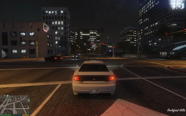 Capture d'écran de gameplay de Grand Theft Auto V montrant une voiture de sport blanche arrêtée à une intersection du quartier de Rockford Hills à Los Santos la nuit, avec les bâtiments de la ville et les feux de circulation en arrière-plan.