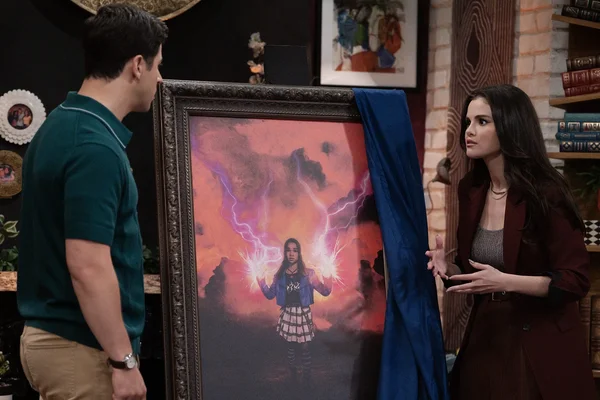 David Henrie et Selena Gomez posant de part et d'autre d'un grand tableau représentant une jeune fille aux mains lumineuses, dans une pièce aux murs de briques et aux bibliothèques.