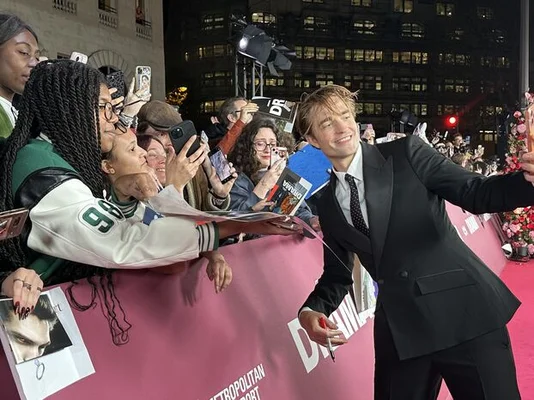 Robert Pattinson en costume noir sur un tapis rose, prenant un selfie avec des fans lors de l'avant-première de The Drama à Paris