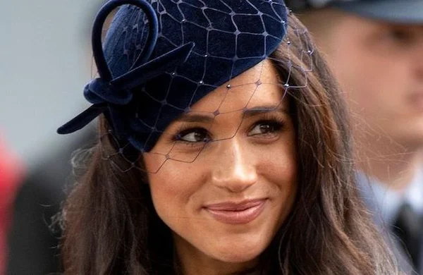 Meghan Markle portant un chapeau fascinator bleu marine avec voile en résille, souriant légèrement, regardant sur le côté.