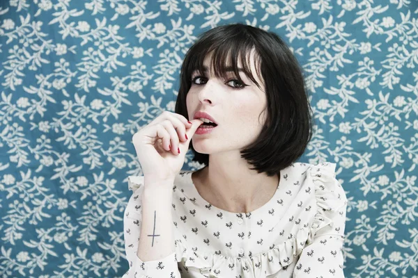 Liza Manili, chanteuse et candidate de Star Academy, pose en chemisier blanc à motifs, avec une coupe au carré sombre et une frange, devant un mur orné de papier peint floral bleu, montrant un tatouage de croix au poignet.