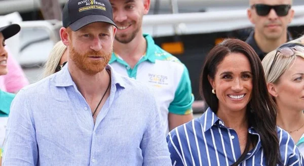 Le prince Harry portant une casquette noire et une chemise bleue, et Meghan Markle en chemisier rayé, debout côte à côte à l'extérieur.