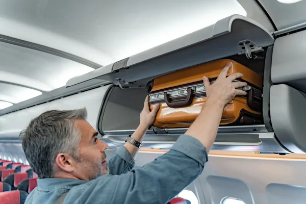 Un homme d'âge mûr en chemise bleue rangeant une valise orange dans le compartiment supérieur d'un avion, illustrant la question des bagages à main en cabine.
