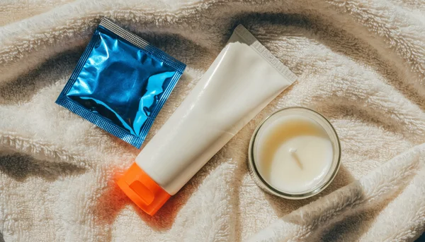 Nature morte éducative : un tube de lubrifiant, un préservatif et une petite bougie posés sur un tissu clair. Illustration douce aux tons pastel évoquant la préparation sereine et la protection.