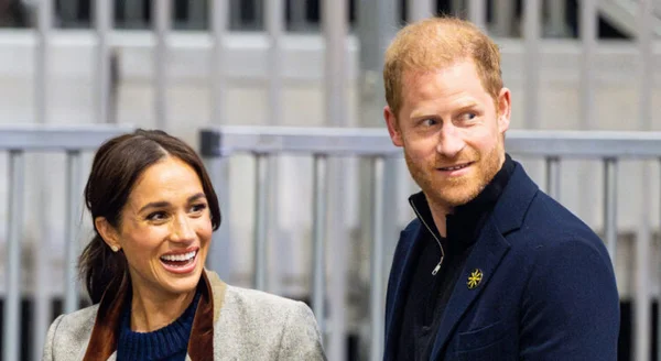 Le prince Harry et Meghan Markle debout côte à côte à l'extérieur, souriant et regardant hors champ vers la droite, barrière métallique floue en arrière-plan.