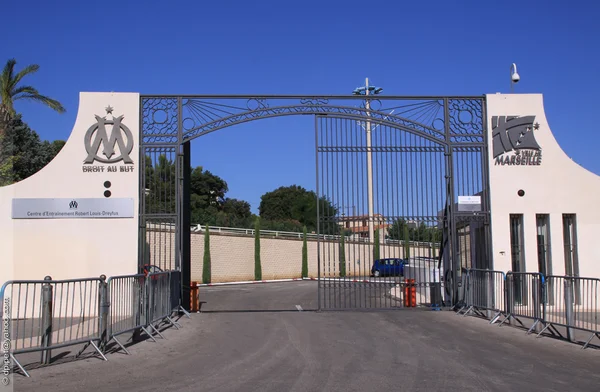 Portail d'entrée du centre d'entraînement Robert-Louis-Dreyfus de l'Olympique de Marseille, avec le logo et la devise du club sur le pilier gauche et l'emblème de la ville de Marseille sur le pilier droit.