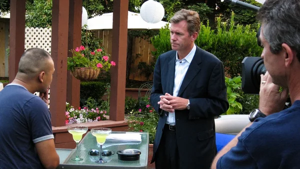 Un homme en costume sombre et chemise bleu clair, debout sur une terrasse, parle à un homme assis pendant qu'un caméraman filme la scène avec une caméra vidéo professionnelle. Le décor comprend des paniers de fleurs suspendus, des lanternes blanches et une table en verre avec deux verres à margarita