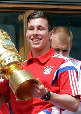 Pierre-Emile Højbjerg, souriant, vêtu d'un polo rouge du Bayern Munich, présentant le trophée de la DFB-Pokal sur le balcon de l'hôtel de ville de Munich, entouré de supporters.