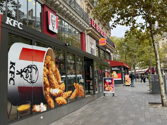 Façade d'un restaurant KFC avec de grandes baies vitrées montrant du poulet frit, situé à côté d'un Burger King sur un trottoir bordé d'arbres.