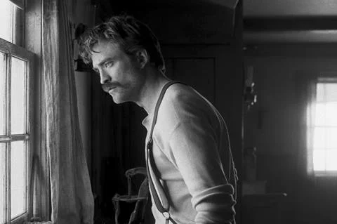 Photographie en noir et blanc de Robert Pattinson avec une moustache, portant des bretelles, de profil regardant par une fenêtre, éclairé de manière dramatique, lors de son rôle dans The Lighthouse.